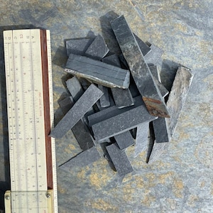 May include: A pile of dark gray slate tiles of various sizes and shapes, next to a ruler marked in inches.