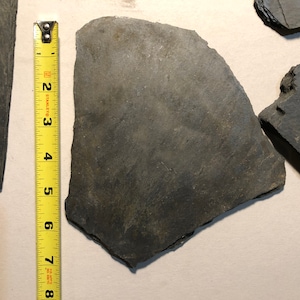 May include: Three pieces of dark gray slate, one of which is measured with a yellow tape measure. The slate is approximately 7 inches long.