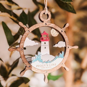 Può includere: Ornamento in legno a forma di timone, con una scena di faro. Il faro ha una parte superiore rossa ed è ambientato su un cielo bianco con nuvole. Il testo "Peggy's Cove Nova Scotia" è visibile.