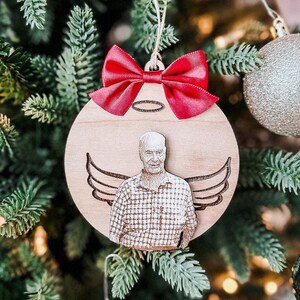 Peut inclure: Une décoration de Noël en bois avec un nœud rouge et une photo d'une personne avec des ailes d'ange. La décoration est accrochée à un sapin de Noël.