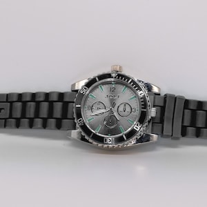 May include: A wristwatch with a silver-toned dial, three sub-dials, and a black rotating bezel. The watch features a silver-toned case and a dark gray, textured silicone strap with a silver-toned buckle. The dial displays the text JBW.