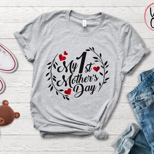 May include: Gray t-shirt with the text "My 1st Mother's Day" in black lettering with a wreath of leaves and red hearts around the text.