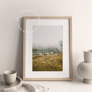 May include: A framed print of a foggy landscape with a herd of sheep grazing on a grassy hillside. The print is in a light wood frame and is displayed on a white surface with a white linen cloth and a ceramic vase.