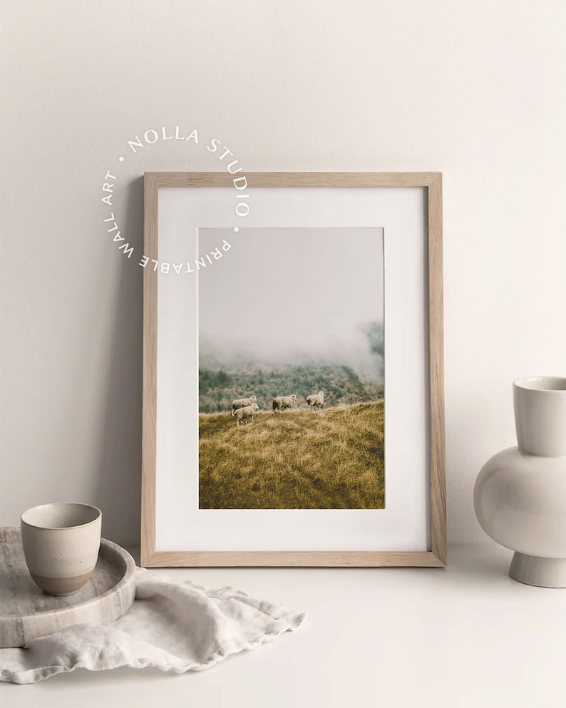 May include: A framed print of a foggy landscape with a herd of sheep grazing on a grassy hillside. The print is in a light wood frame and is displayed on a white surface with a white linen cloth and a ceramic vase.