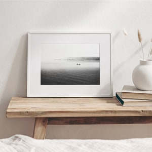 May include: A black and white photograph of a boat on a lake with a city skyline in the distance. The photo is framed in a white frame and is sitting on a wooden shelf.