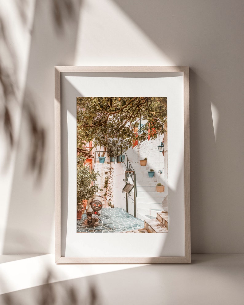 May include: A framed photograph of a narrow, white-walled alleyway in a European city. The alleyway is lined with potted plants and a scooter is parked at the bottom of the steps.