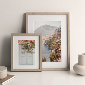 May include: Two framed photographs, one with a view of a coastal town and the other with a white door with pink flowers. Both frames are light brown wood.