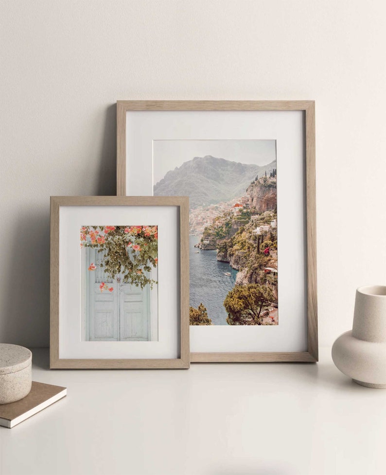 May include: Two framed photographs, one with a view of a coastal town and the other with a white door with pink flowers. Both frames are light brown wood.