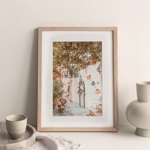 May include: A framed photograph of a white stone staircase with potted plants and a small scooter in a narrow alleyway. The photo is in a light wood frame and is sitting on a white surface.