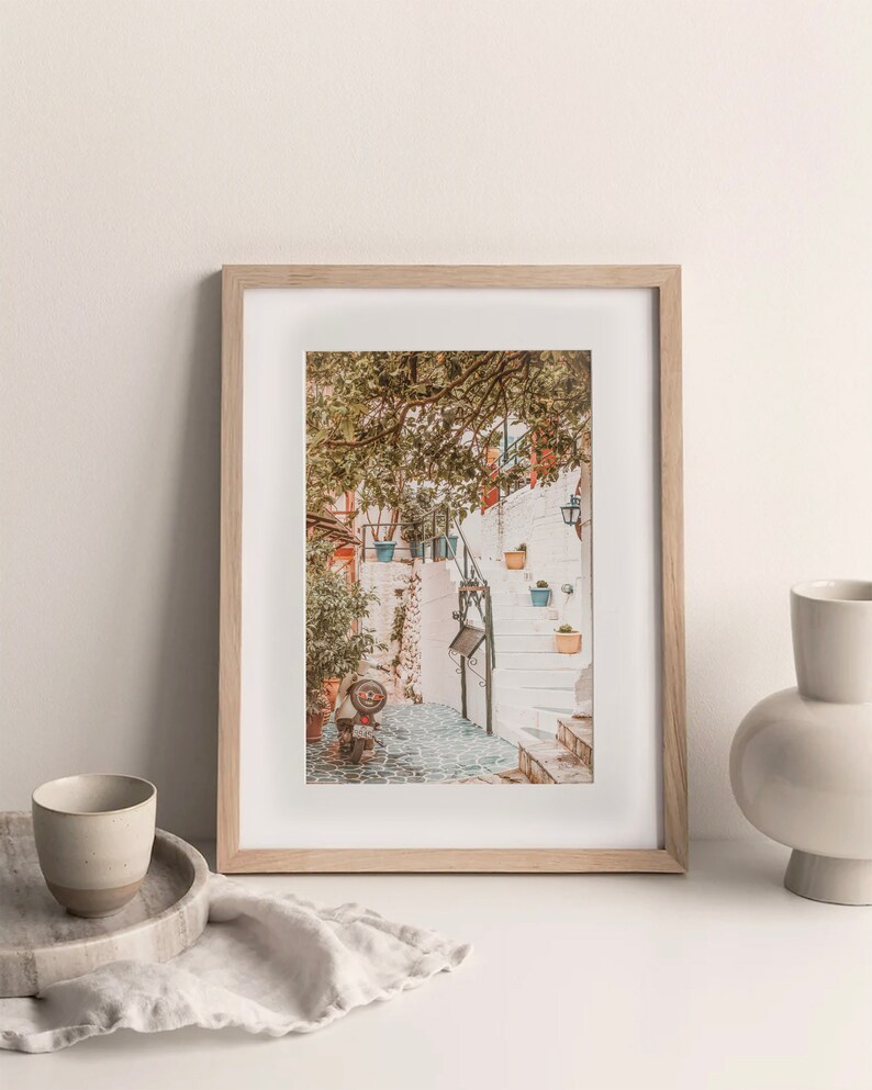 May include: A framed photograph of a white stone staircase with potted plants and a small scooter in a narrow alleyway. The photo is in a light wood frame and is sitting on a white surface.