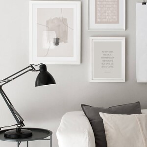May include: Three framed prints on a white wall above a white sofa with grey and white cushions. The prints are all in white frames. The left print is a minimalist abstract design with brown and grey shapes. The middle print is a pattern of white lines on a light pink background. The right print is a quote that reads "You don't always need a plan. Sometimes you just need to breathe. Trust, let go, and see what happens."