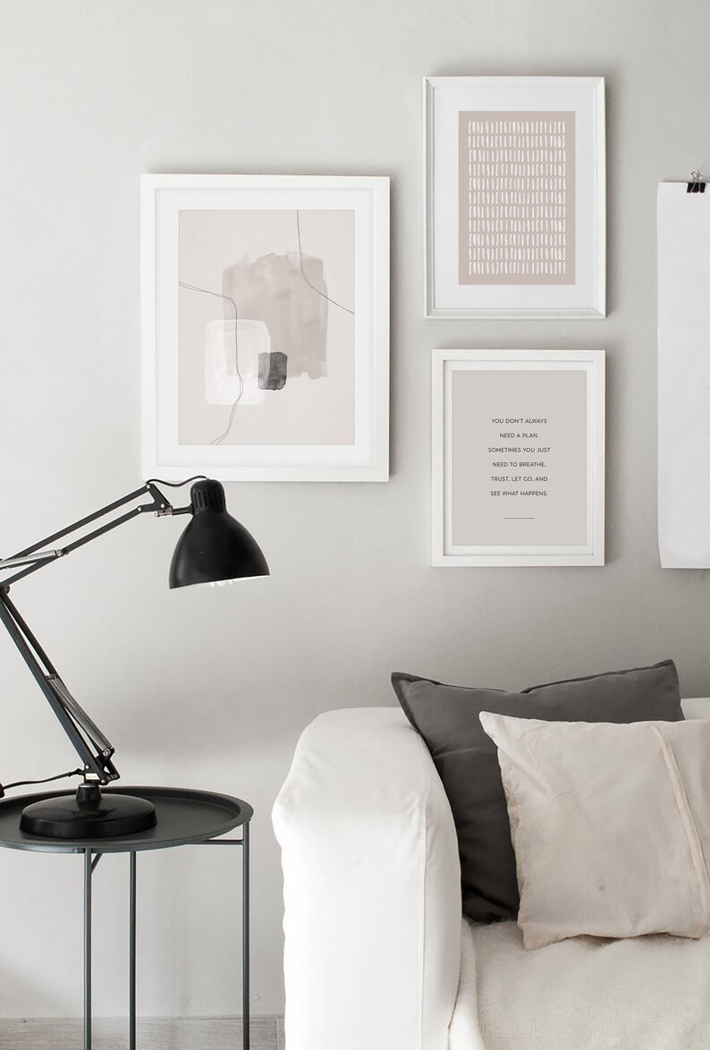 May include: Three framed prints on a white wall above a white sofa with grey and white cushions. The prints are all in white frames. The left print is a minimalist abstract design with brown and grey shapes. The middle print is a pattern of white lines on a light pink background. The right print is a quote that reads "You don't always need a plan. Sometimes you just need to breathe. Trust, let go, and see what happens."