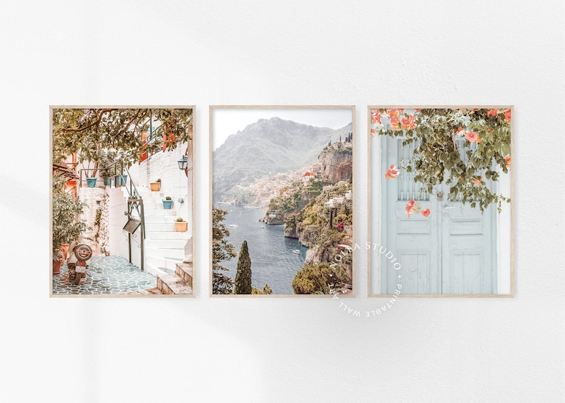 May include: Set of three printable wall art prints featuring coastal Italian scenery. The first print shows a white building with a stairway and lush greenery. The second print shows a view of a town on a cliffside overlooking a blue body of water. The third print shows a white door with pink flowers and green vines.