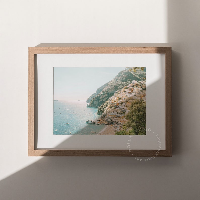 May include: A framed photograph of a coastal town built into a hillside, with the ocean in the foreground. The town is a mix of white and yellow buildings, with green trees and foliage. The sky is a light blue, and the water is a turquoise blue.