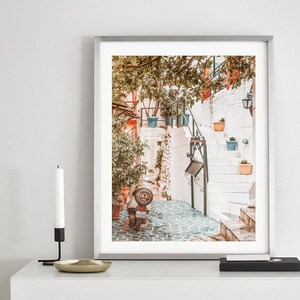 May include: A vintage scooter parked in a narrow, cobblestone alleyway in a Mediterranean town. The alleyway is lined with whitewashed buildings and potted plants. The scooter is facing away from the camera, and the back tire is visible.