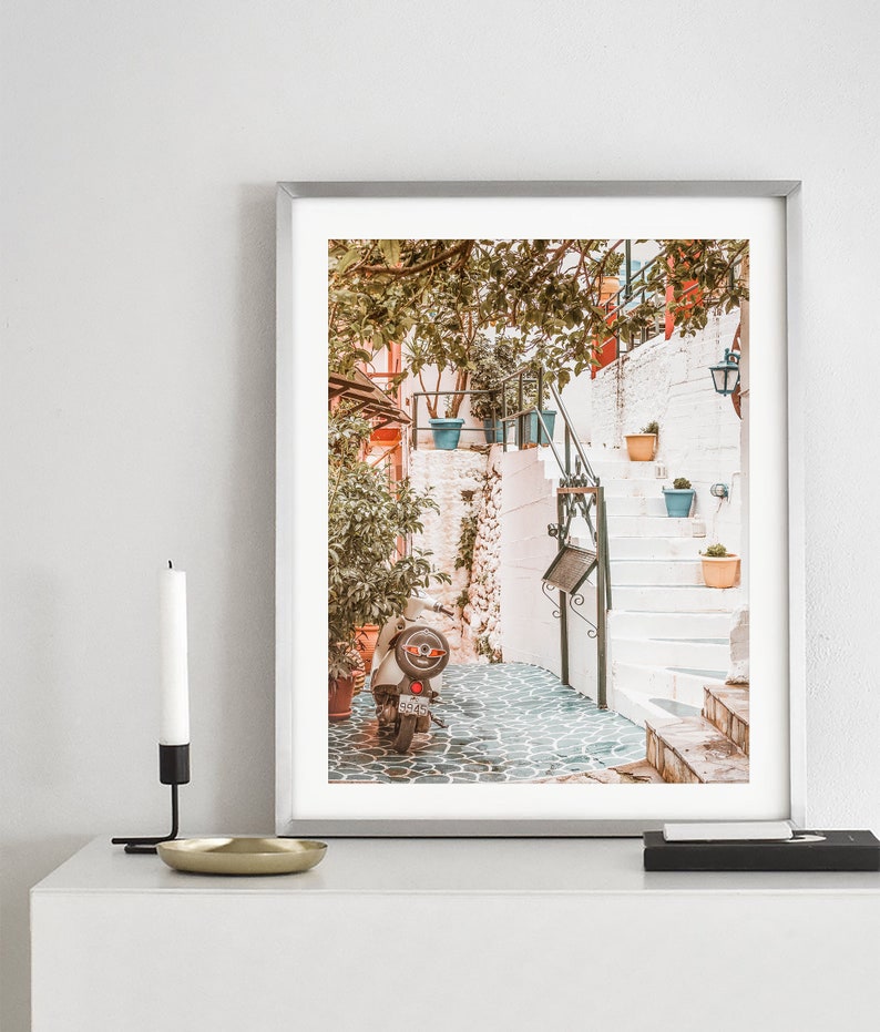 May include: A vintage scooter parked in a narrow, cobblestone alleyway in a Mediterranean town. The alleyway is lined with whitewashed buildings and potted plants. The scooter is facing away from the camera, and the back tire is visible.