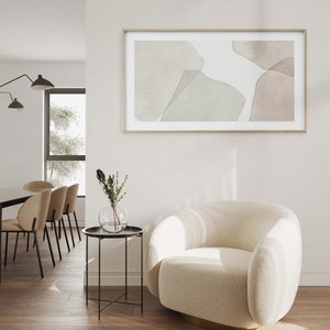 Op de afbeelding: Een witte, pluche fauteuil met een gouden basis staat in een woonkamer met een bijzettafel en een ingelijst abstract schilderij aan de muur. Het schilderij heeft zachte, gedempte kleuren in bruin en groen.