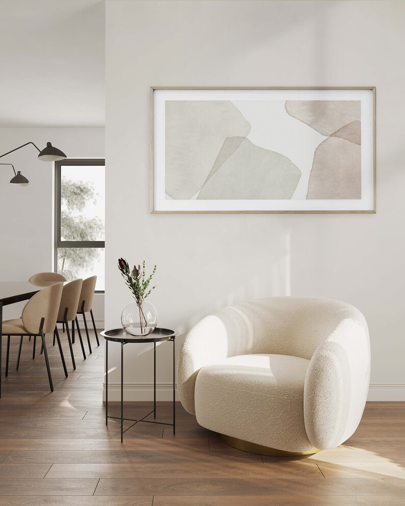 Op de afbeelding: Een witte, pluche fauteuil met een gouden basis staat in een woonkamer met een bijzettafel en een ingelijst abstract schilderij aan de muur. Het schilderij heeft zachte, gedempte kleuren in bruin en groen.