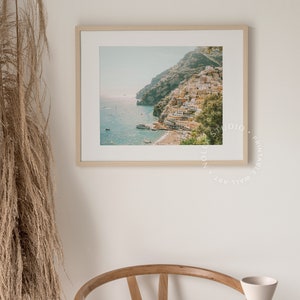 May include: A framed photograph of a coastal town built into a hillside, with a blue ocean in the foreground. The town is a mix of white and yellow buildings, with green trees and foliage.