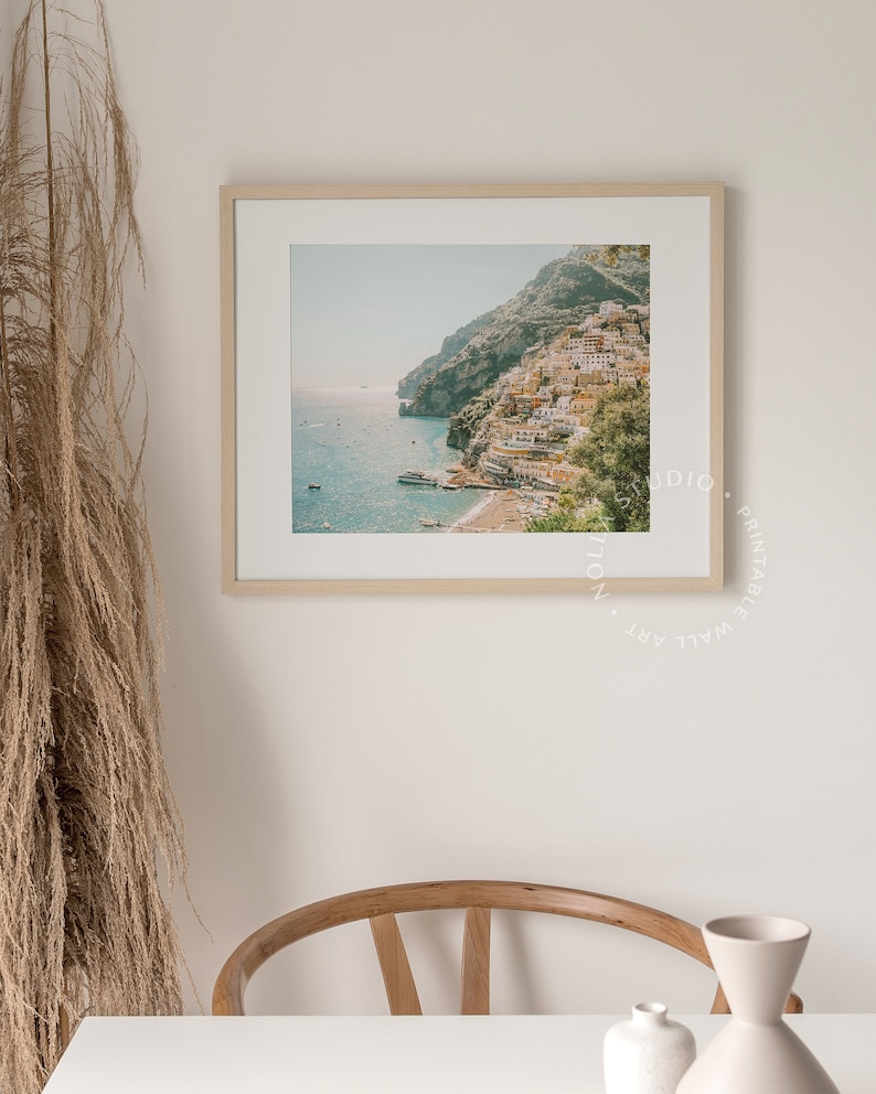 May include: A framed photograph of a coastal town built into a hillside, with a blue ocean in the foreground. The town is a mix of white and yellow buildings, with green trees and foliage.