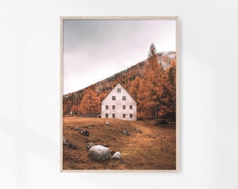 May include: A white house with multiple windows sits on a grassy hill surrounded by trees with orange and yellow leaves. The sky is overcast and the ground is covered in fallen leaves.