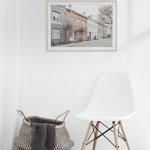 May include: A framed photograph of a row of colorful houses with a tree in front of them. The houses are in a row and have a sidewalk in front of them. There is a white chair and a woven basket in the foreground.