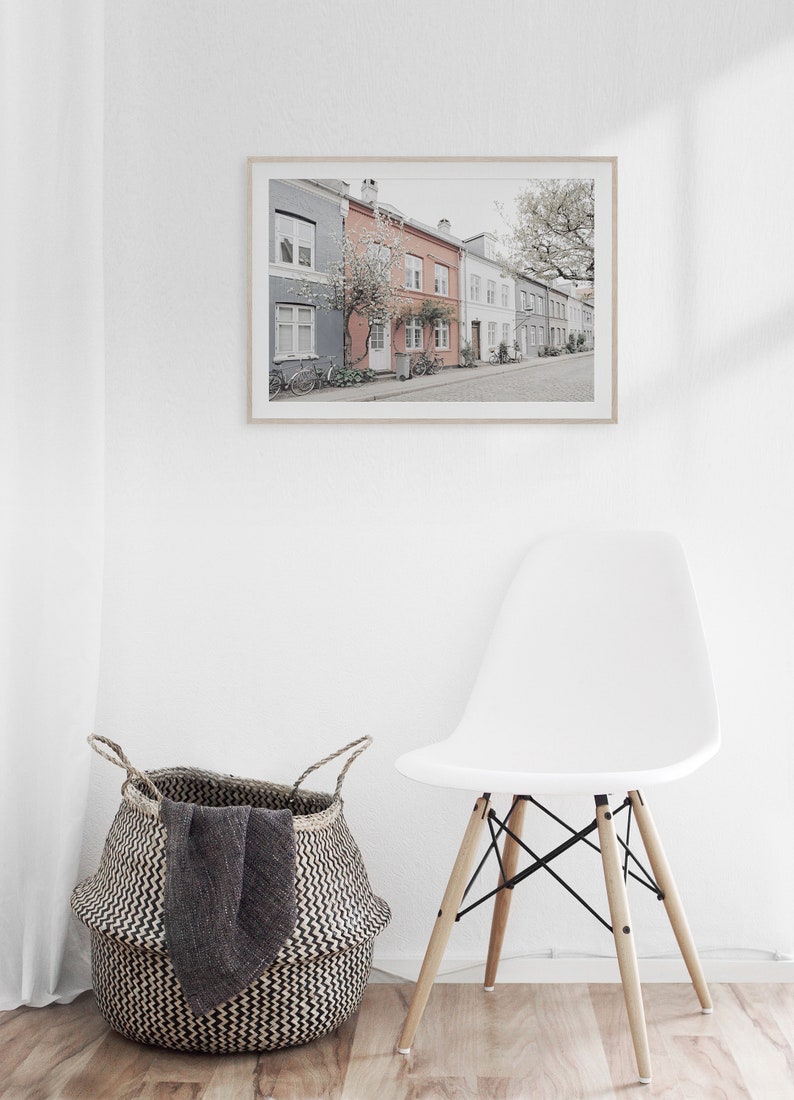 May include: A framed photograph of a row of colorful houses with a tree in front of them. The houses are in a row and have a sidewalk in front of them. There is a white chair and a woven basket in the foreground.