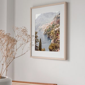 May include: A framed photograph of a coastal town nestled on a hillside overlooking a blue body of water. The town is surrounded by lush green vegetation and the water is dotted with boats.