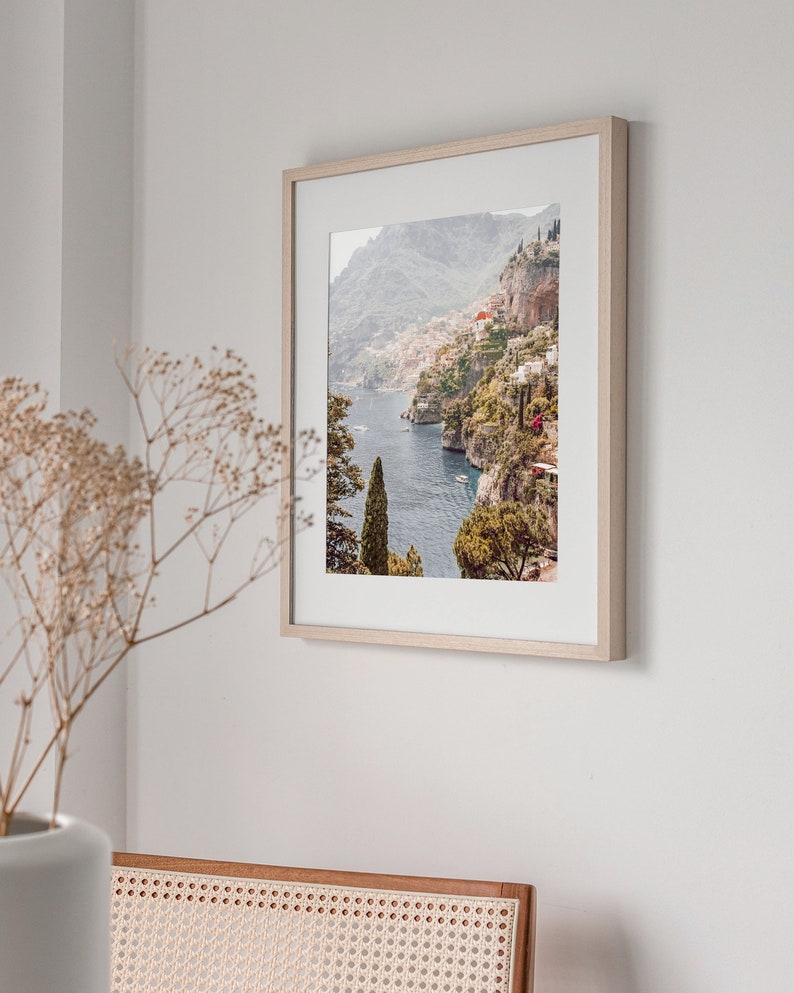 May include: A framed photograph of a coastal town nestled on a hillside overlooking a blue body of water. The town is surrounded by lush green vegetation and the water is dotted with boats.