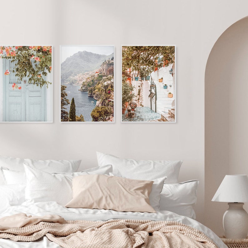 May include: Three framed prints of Italian coastal scenes. The first print shows a blue door with a flowering vine. The second print shows a view of a coastal town with a mountain in the background. The third print shows a white building with a staircase and potted plants.