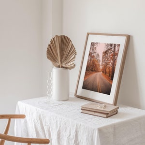 May include: A framed photograph of a road through a forest with fall foliage. The photo is on a white table with a vase and a dried palm leaf.