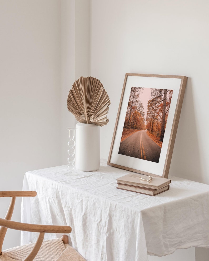May include: A framed photograph of a road through a forest with fall foliage. The photo is on a white table with a vase and a dried palm leaf.