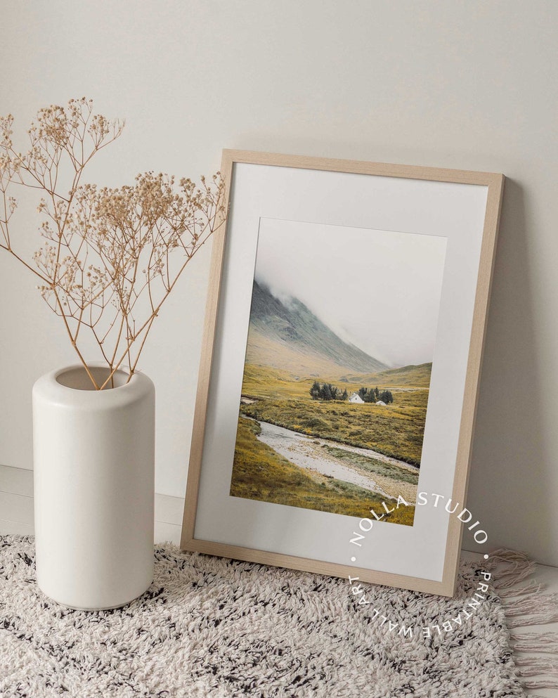 May include: A framed print of a landscape photograph with a mountain range, a river, and a small house in the distance. The print is in a light wood frame and has a white mat. The print is on a white rug with a vase of dried flowers.