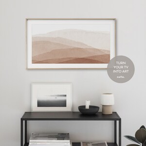 May include: A modern living room interior with a large framed abstract painting of brown and white watercolor mountains hanging on a white wall. A smaller black and white photo of a lake with two boats is on a black shelf below the painting. A white ceramic lamp and a black bowl with a candle are on the shelf. The text "TURN YOUR TV INTO ART" is on a white circle with a black outline. The text "nolla." is below the circle.