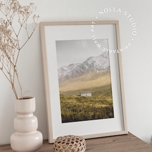 May include: A framed print of a landscape photograph featuring a small house in a field with snow-capped mountains in the background. The print is in a light wood frame and is sitting on a wooden table with a vase of dried flowers and a woven basket.