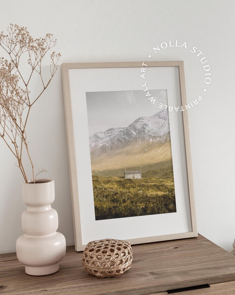 May include: A framed print of a landscape photograph featuring a small house in a field with snow-capped mountains in the background. The print is in a light wood frame and is sitting on a wooden table with a vase of dried flowers and a woven basket.