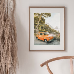 May include: A vintage orange Fiat car driving on a winding road with a view of the ocean and green hills in the background.