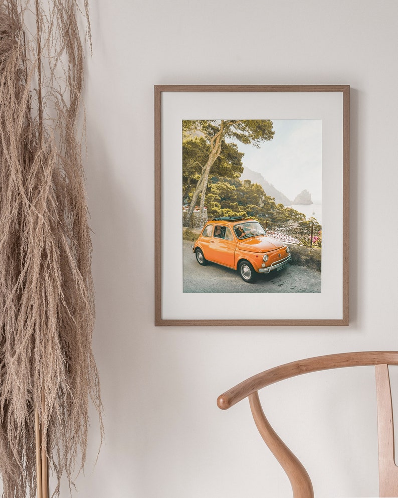 May include: A vintage orange Fiat car driving on a winding road with a view of the ocean and green hills in the background.
