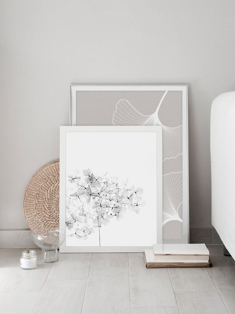 May include: Two framed black and white botanical prints, one with a ginkgo leaf design and the other with a delicate flower design, are displayed on a white wall. The prints are leaning against a white bed frame.