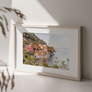 Pu&ograve; includere: Una fotografia incorniciata di un pittoresco villaggio italiano arroccato su una collina che domina un mare blu. Le case sono dipinte in tonalit&agrave; di giallo, arancione e rosso, e il paesaggio circostante &egrave; lussureggiante e verde.