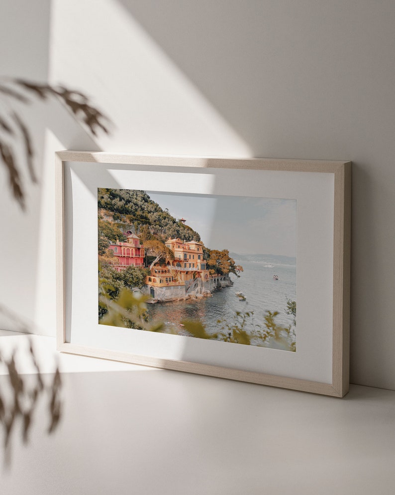Pu&ograve; includere: Una fotografia incorniciata di un pittoresco villaggio italiano arroccato su una collina che domina un mare blu. Le case sono dipinte in tonalit&agrave; di giallo, arancione e rosso, e il paesaggio circostante &egrave; lussureggiante e verde.