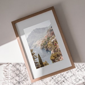 May include: A framed photograph of a coastal landscape with a mountain range, a blue body of water, and lush green trees. The frame is made of light brown wood.