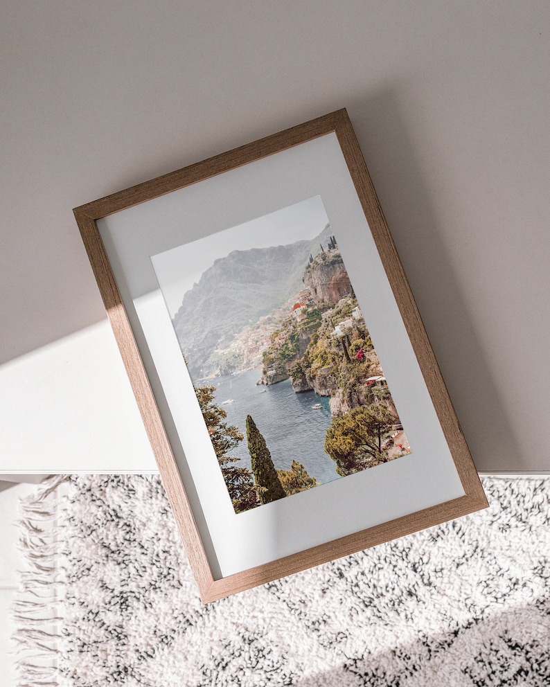 May include: A framed photograph of a coastal landscape with a mountain range, a blue body of water, and lush green trees. The frame is made of light brown wood.