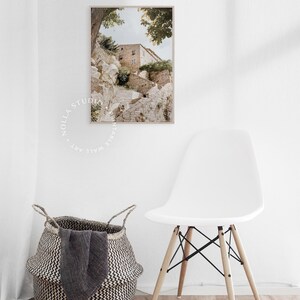 May include: A photograph of a stone building with a staircase leading up to it. The building is surrounded by trees and greenery. The photo is in a light brown frame and is hanging on a white wall.