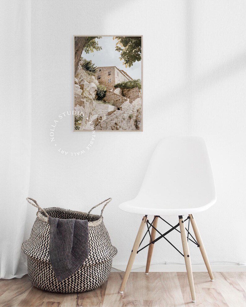 May include: A photograph of a stone building with a staircase leading up to it. The building is surrounded by trees and greenery. The photo is in a light brown frame and is hanging on a white wall.