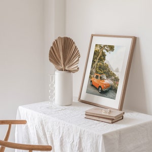 May include: A framed print of an orange vintage car parked on a road with lush green trees and a view of a rocky coastline in the background. The print is on a white table with a dried palm leaf in a white vase and two books.