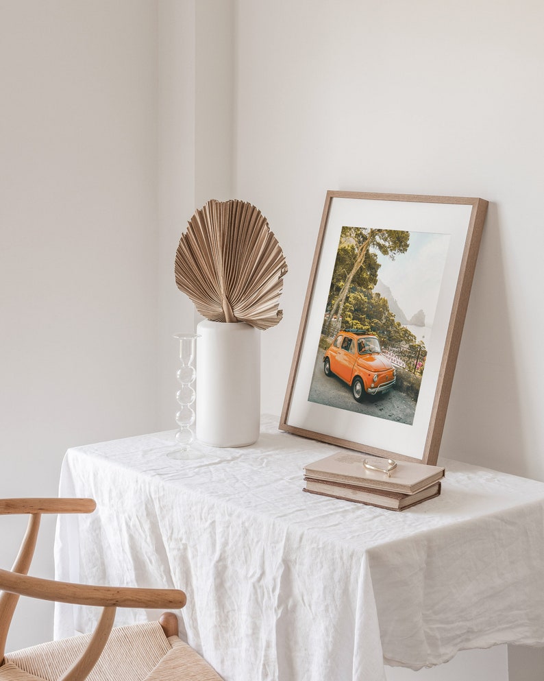 May include: A framed print of an orange vintage car parked on a road with lush green trees and a view of a rocky coastline in the background. The print is on a white table with a dried palm leaf in a white vase and two books.