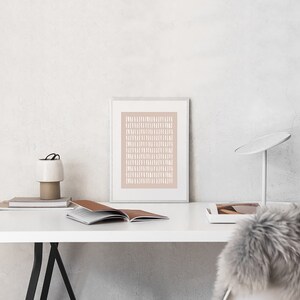 May include: A framed print with a white geometric pattern on a light pink background. The print is hanging on a white wall above a white desk with a lamp, books, and a white chair.