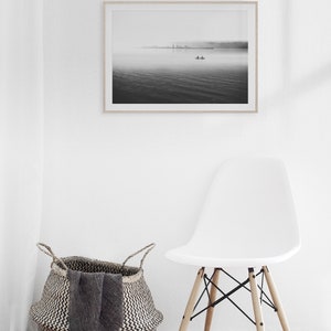 May include: A black and white photograph of a lake with a small boat in the distance. The photo is framed and hanging on a white wall above a white chair and a woven basket.