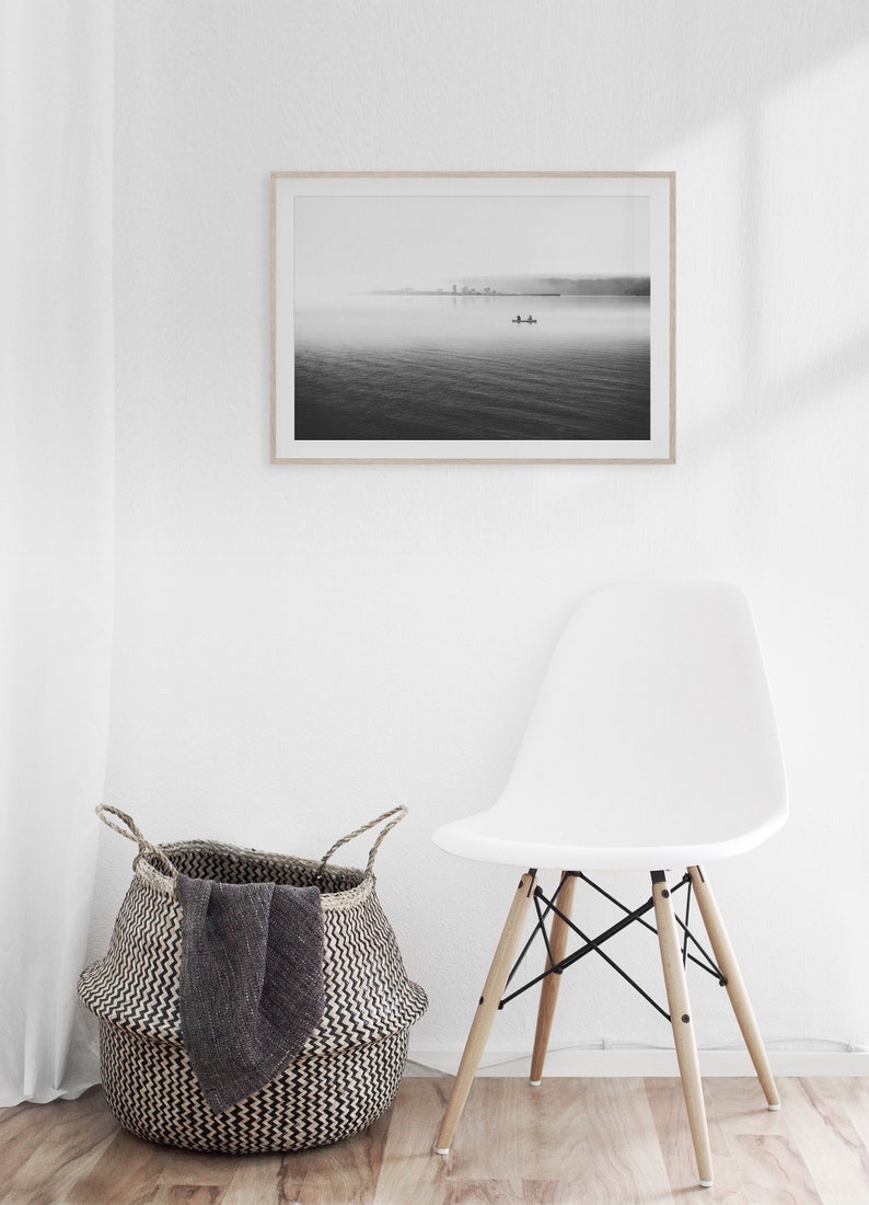May include: A black and white photograph of a lake with a small boat in the distance. The photo is framed and hanging on a white wall above a white chair and a woven basket.